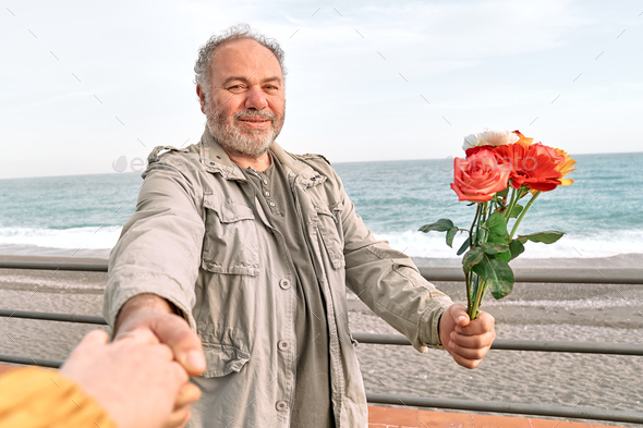 Smiling mature bearded man with colorful flowers holds and pulls hand leading her by the sea. - Stock Photo - Images