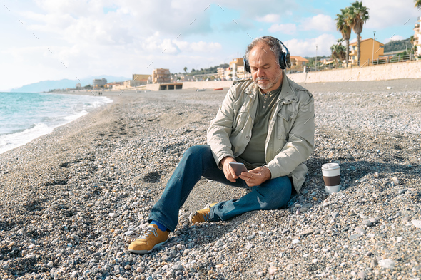 Happy middle-aged bearded man listening music in headphones while sitting on winter beach. - Stock Photo - Images
