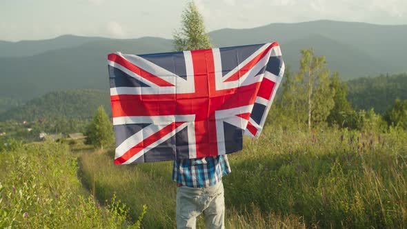 Joyful Man Standing with Big Flag of Great Britain on Top of Mountain at Sunset alt