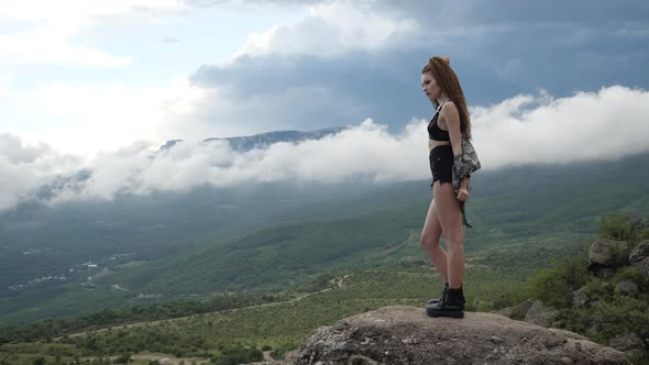 Sexy Girl in Short Shorts and Sporting Black Shoes Stands on Top of the Mountain