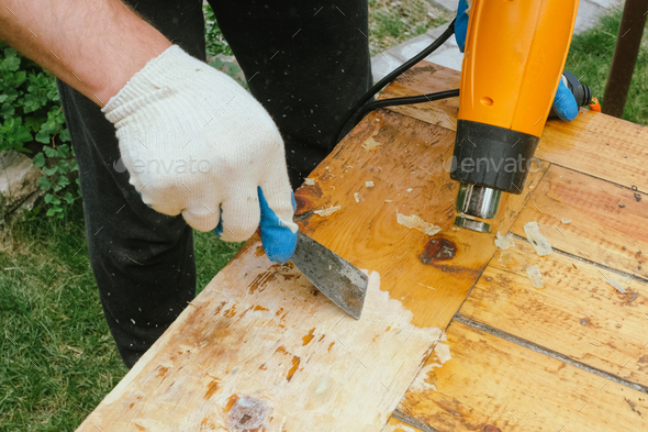 Man removing old varnish from wood using scraper and heat gun.
