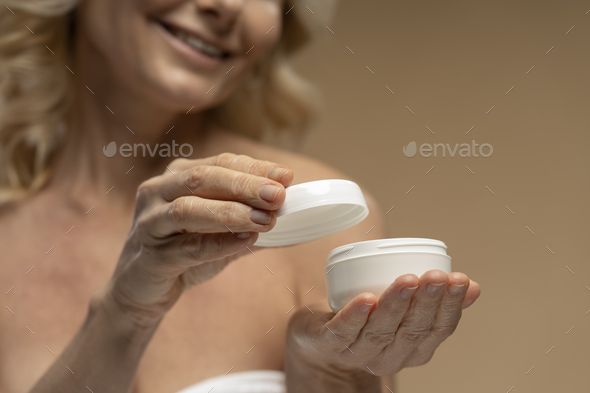 Focus on hands of blurry mature woman opening jar of moisturizing cosmetic product, beige background