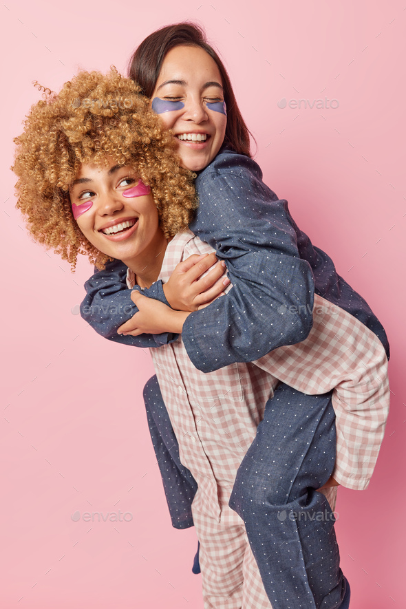 Happy positive young women give piggyback ride dressed in comfortable pajama smile positively have