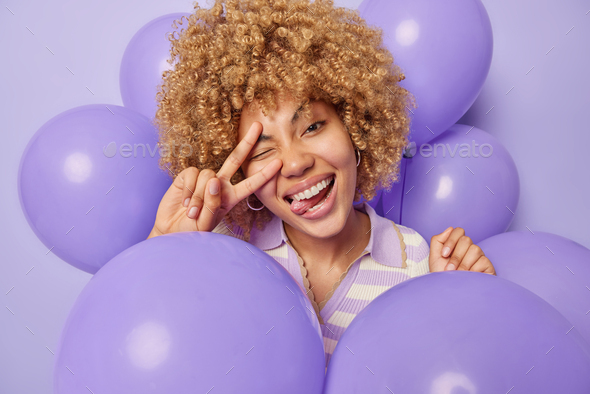 Positive curly haired woman makes peace gesture over eye sticks out tongue has upbeat mood