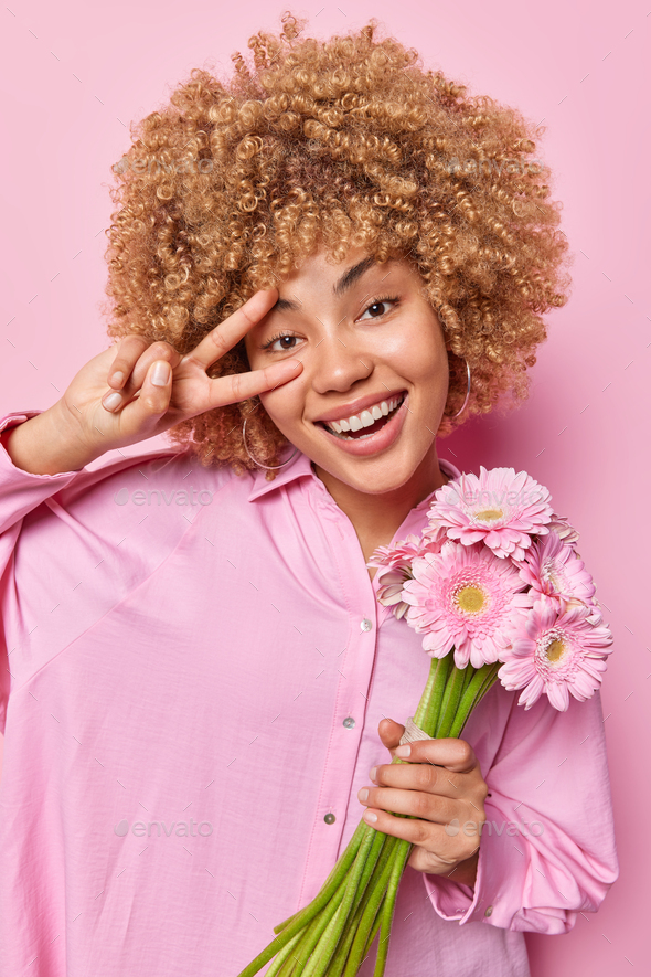 Positive carefree woman with curly hair makes peace sign over eye smiles broadly holds bouquet of