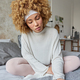 Serious curly haired young woman dressed in casual clothes poses on comfortable bed in bedroom holds - PhotoDune Item for Sale
