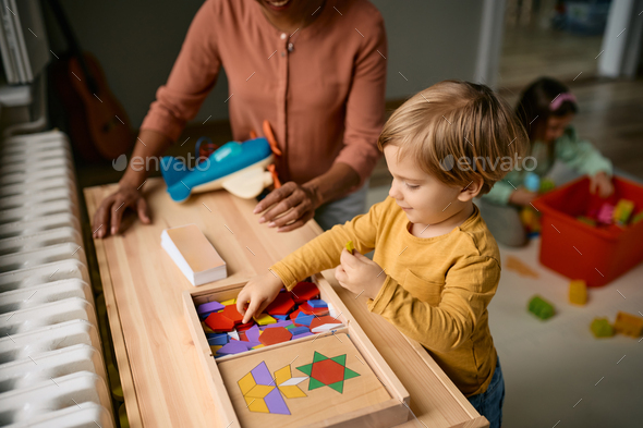 Small boy playing with wooden puzzle shapes with his teacher at kindergarten. - Stock Photo - Images