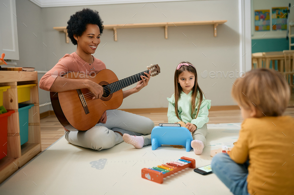Happy black kindergarten teacher playing acoustic guitar to group of kids. - Stock Photo - Images