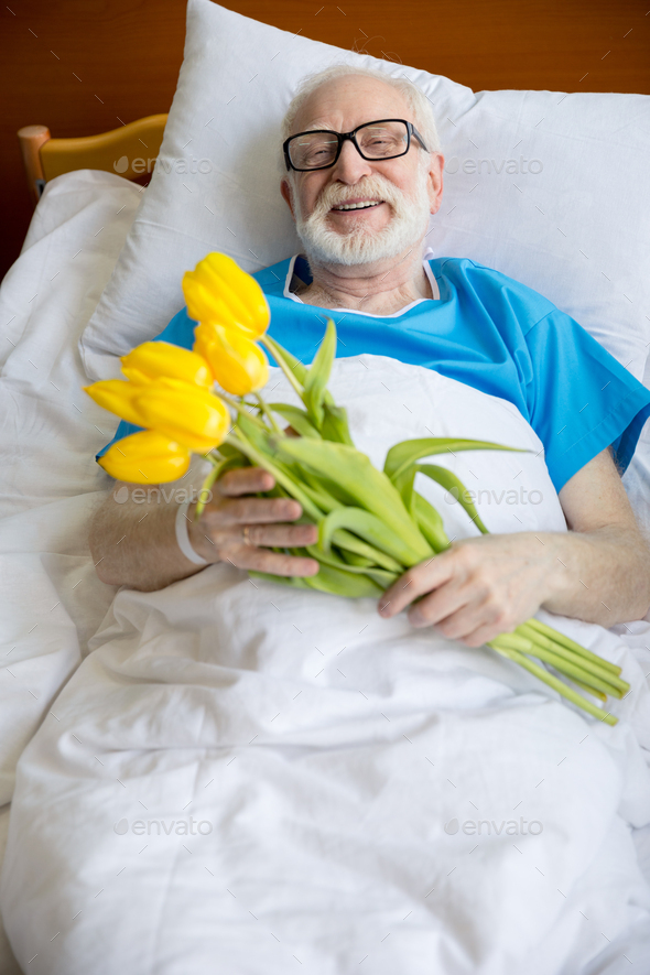 senior male patient in hospital bed with tulip flowers in clinic Stock