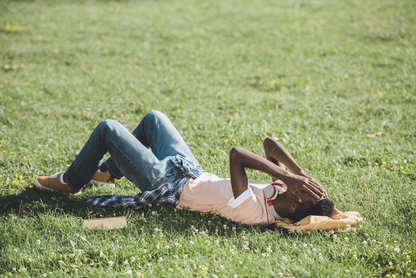 side view of african american student with hands on face resting on green grass in park - Stock Photo - Images