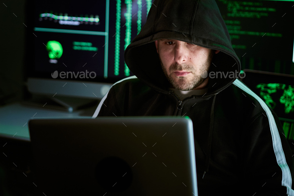 Portrait of criminal hooded hacker at desk and breaking into government or big company data servers