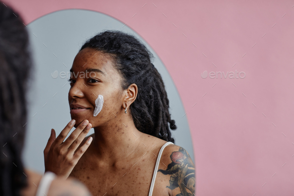 Young black woman with acne scars using face cream looking in mirror - Stock Photo - Images