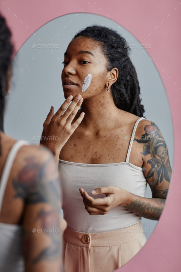 Candid young black woman with acne scars using face cream by mirror - Stock Photo - Images