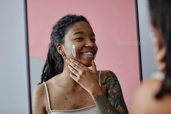Candid smiling black woman with acne scars using face cream by mirror - Stock Photo - Images