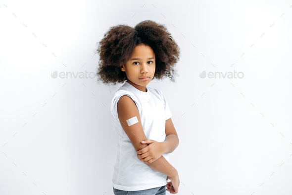 Upset African American curly haired little girl, with a band-aid on her shoulder after vaccination