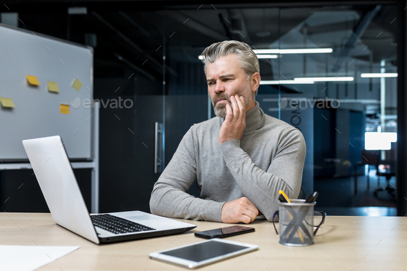 A senior gray-haired man sits in the office, holding his cheek. feels a toothache and discomfort - Stock Photo - Images