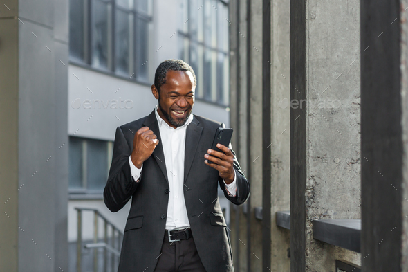 Happy young african american man near office. He holds the phone in his hands, celebrates, rejoices - Stock Photo - Images