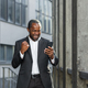Happy young african american man near office. He holds the phone in his hands, celebrates, rejoices - PhotoDune Item for Sale