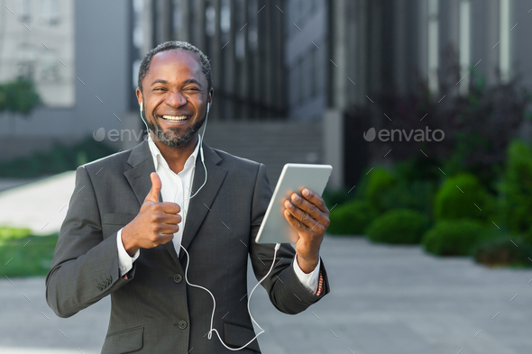 A young African American man in a suit is standing outside wearing headphones, holding a tablet - Stock Photo - Images