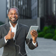 A young African American man in a suit is standing outside wearing headphones, holding a tablet - PhotoDune Item for Sale
