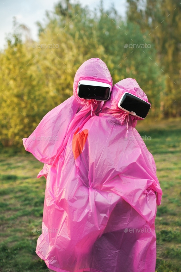 Vertical shot of two people in pink plastic raincoats and VR headsets hugging each other