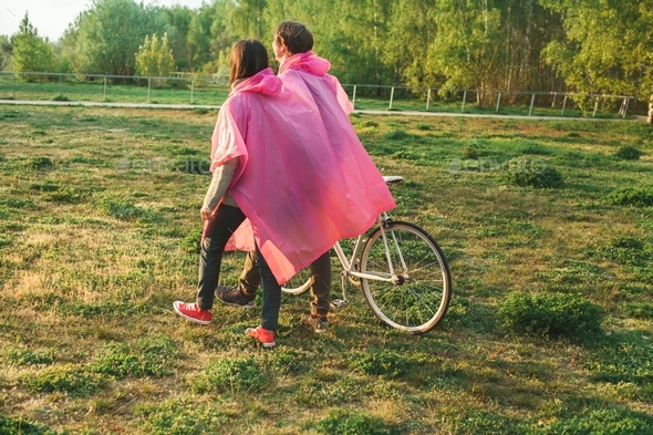 Male and a female in pink plastic raincoats walking in the field with a bicycle on a date