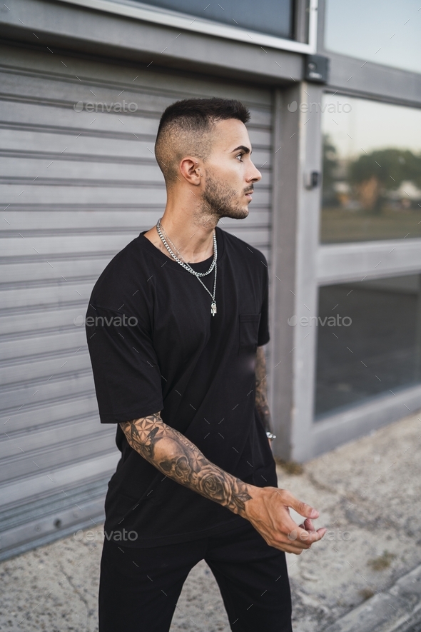 European tattooed hip male in a black sporty outfit and a metal chain  standing in front of a garage Stock Photo by wirestock