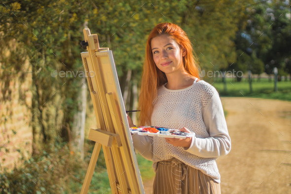 Sunny shot of a redhead female painting on a painting board with palettes in a park