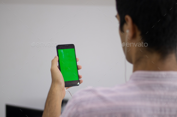 Indian male with headphones in his ears and looking at the green screen of his cellphone