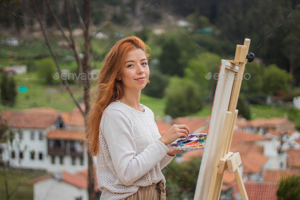 Beautiful shot of a redhead female painting on a painting board with palettes in a park