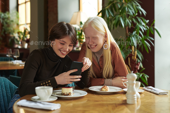 Two young cheerful girlfriends discussing online photo or video in mobile phone - Stock Photo - Images