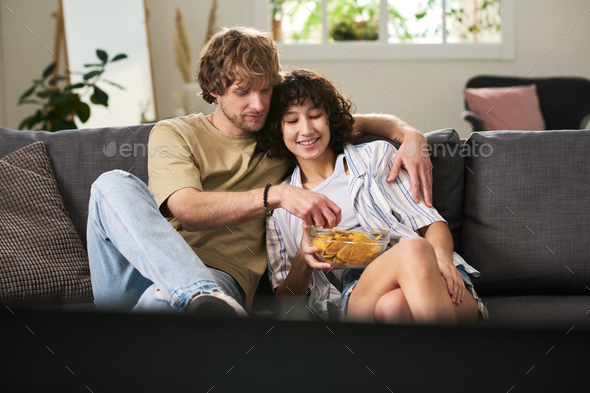Young man taking potato chips from bowl held by his wife