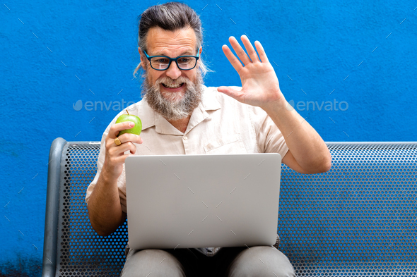 Mature caucasian man eating apple waving hello on a video call in park bench.