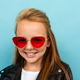 Caucasian teenager girl with brown hair in black jacket holds red sunglasses isolated on blue - PhotoDune Item for Sale