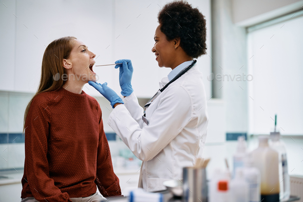 African American doctor using tongue depressor while checking woman\'s throat at medical clinic.