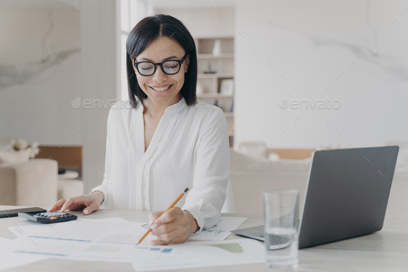 Smiling businesswoman in glasses works calculating project budget sitting at office desk with laptop - Stock Photo - Images