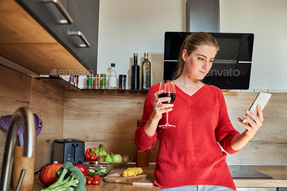 A charming young woman stands in the kitchen and is distracted from cooking using a mobile phone.