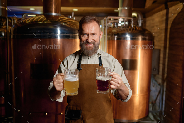 Satisfied senior brewer posing for camera with pale ale and dark beer pint