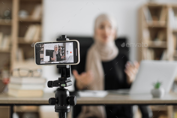 Blur background of pretty muslim woman with headscarf sitting at desk and filming video blog.