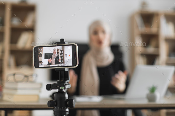 Blur background of pretty muslim woman with headscarf sitting at desk and filming video blog.