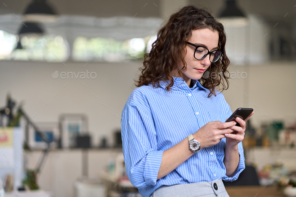 Young business woman holding mobile phone standing in office using cell. - Stock Photo - Images