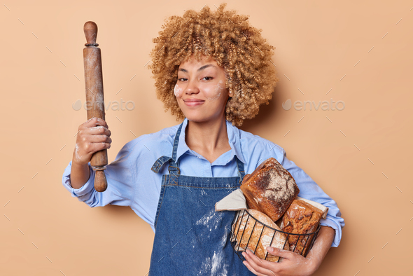 Boastful female baker holds basket of delicious fresh bread and wooden rolling pin demonstrates her - Stock Photo - Images