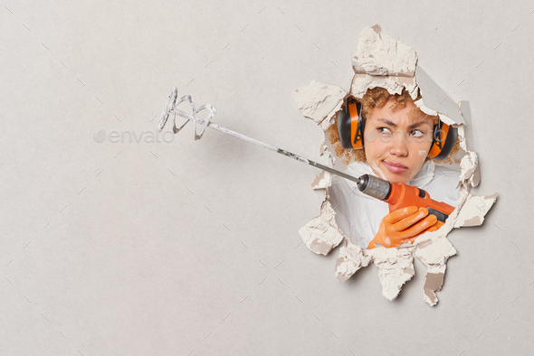 Unhappy serious female construction worker holds mixer busy with home repair wears headphones for - Stock Photo - Images