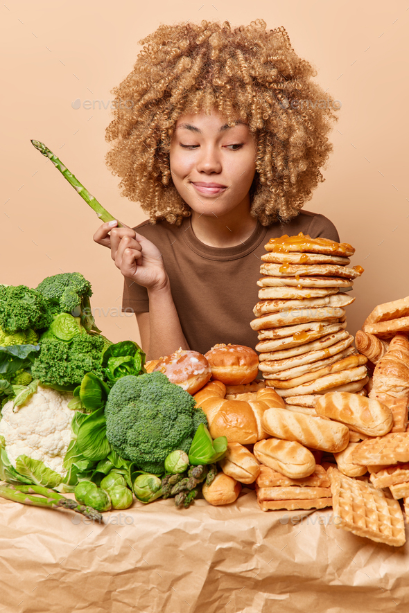 Vertical shot of serious curly haired woman holds asparagus looks with temptation at delicious sweet - Stock Photo - Images