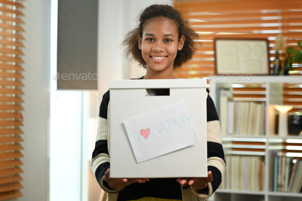 Smiling young African woman holding box of clothes with donate label.