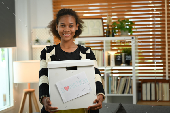 Portrait of smiling young woman holding box of clothes with donate label.
