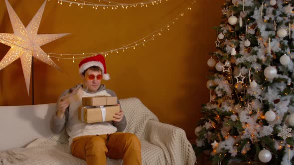 Man in Santa Hat and Bright Glasses with Gift Boxes Sitting on Couch Near Christmas Tree alt