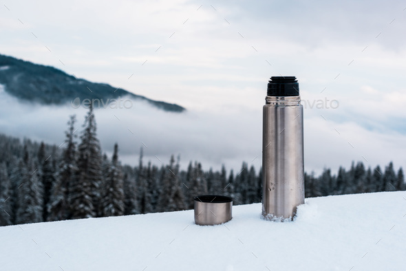 open metallic vacuum flask in snowy mountains with pine trees and white fluffy clouds