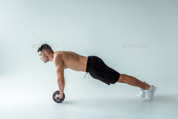 side view of sexy muscular bodybuilder with bare torso exercising with ab wheel on grey background