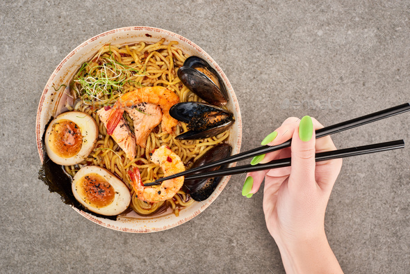 cropped view of woman holding chopsticks near spicy seafood ramen on grey surface - Stock Photo - Images